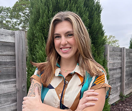 A photo of STUN employee Rachel, smiling standing in front of a green tree.
