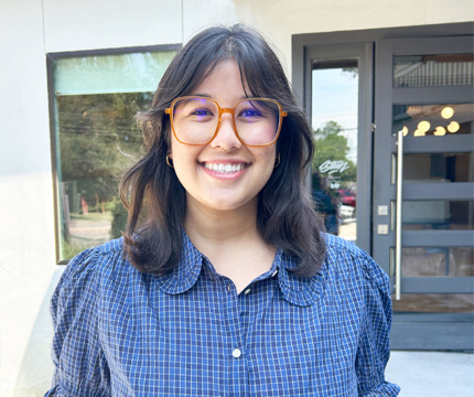 Smiling woman with yellow-framed glasses and a blue checkered shirt standing outside a modern building.