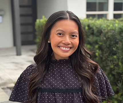 Portrait of a smiling woman with long dark hair in a black polka-dot blouse outdoors with greenery behind her.